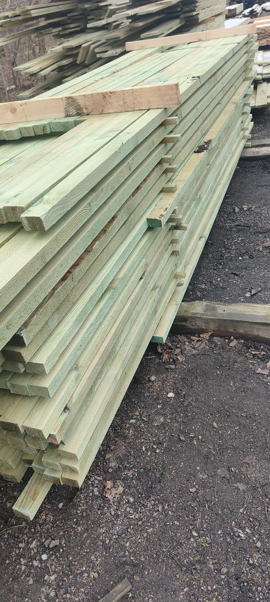 Stos impregnowanej tarcicy budowlanej ułożony na ziemi, widoczne sęki i zielonkawy odcień impregnatu.
