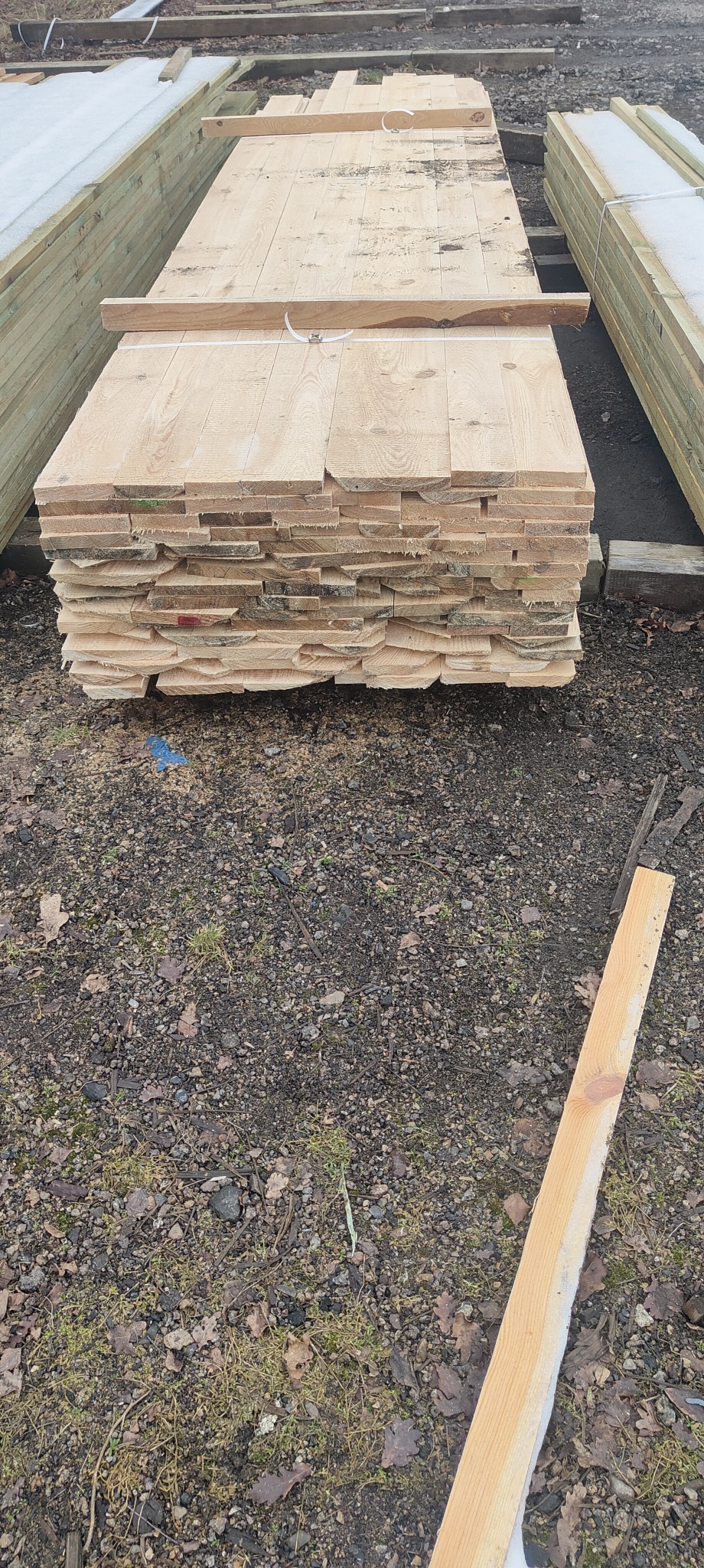 Stos jasnej tarcicy budowlanej z widocznymi krawędziami, ułożony na żwirowym podłożu. W tle widoczne inne pakiety drewna, niektóre pokryte śniegiem.