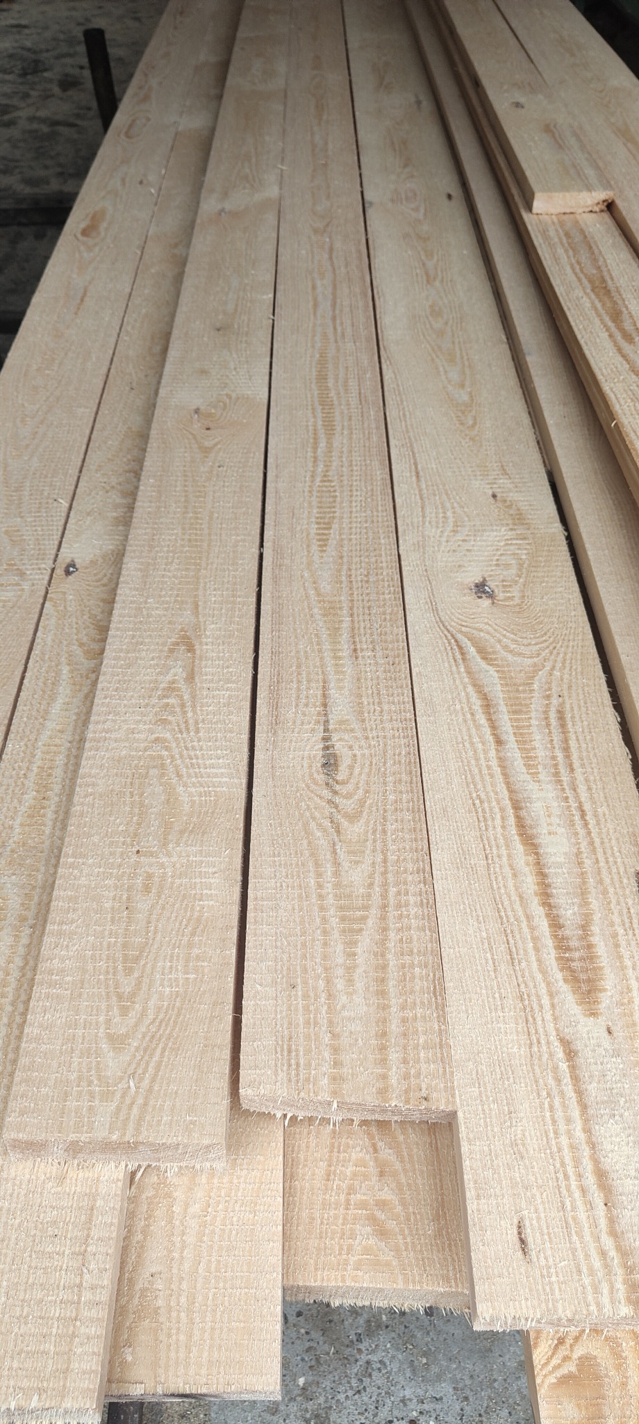 Stos jasnych, świeżo przetartych desek z widocznym usłojeniem, ułożonych warstwami na szarym podłożu, z niewielkimi fragmentami kory na krawędziach niektórych elementów.