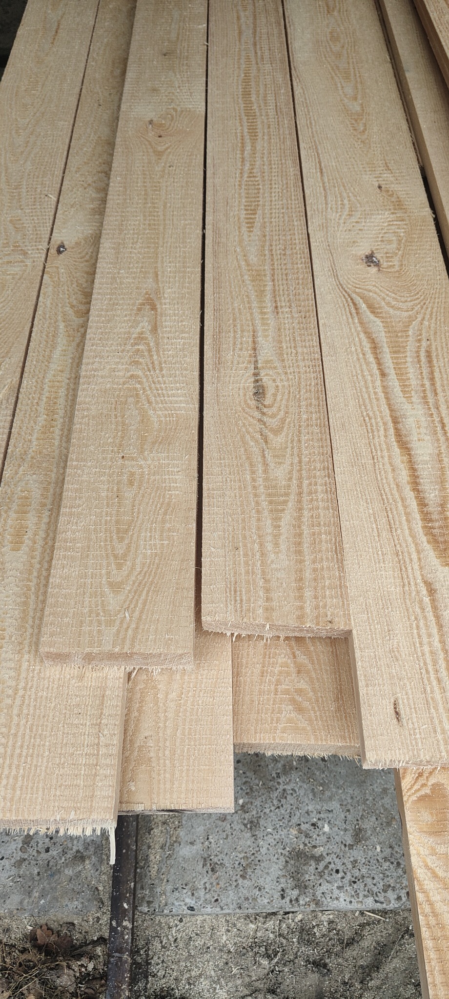 Stos jasnej, świeżo przetartej tarcicy sosnowej z widocznym usłojeniem, ułożonej na szorstkiej, betonowej posadzce.