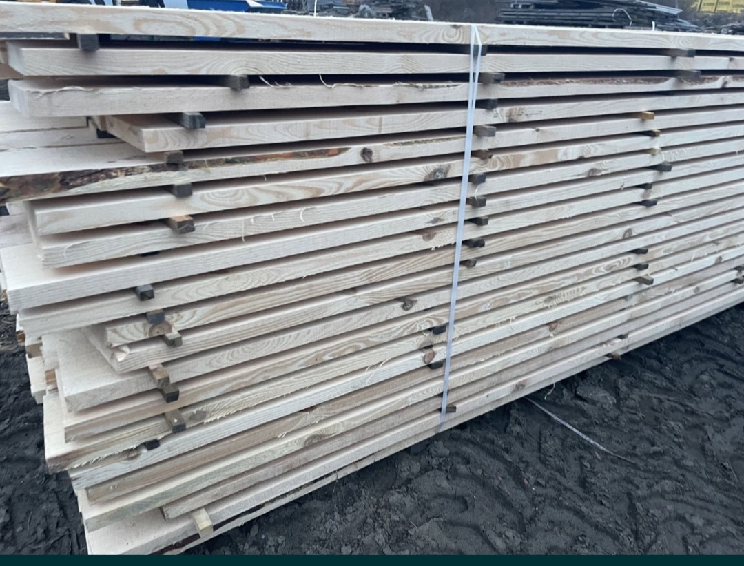 Stos jasnych desek budowlanych związanych białą taśmą, widoczne naturalne krawędzie z korą, ułożone na ciemnym, błotnistym podłożu.