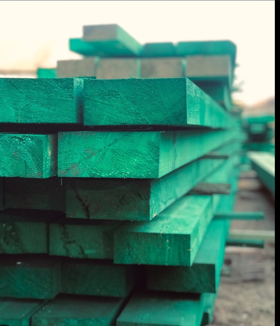 Stos impregnowanych na zielono kantówek z widocznym rysunkiem słojów drewna, ułożonych równo na placu składowym.