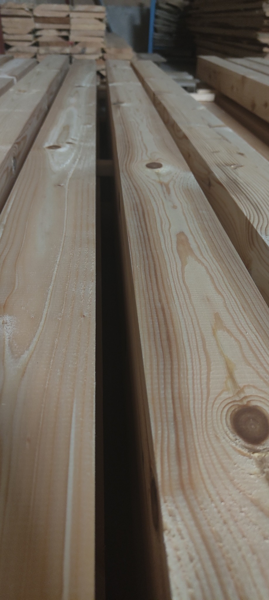 Ujęcie z bliska na stos jasnego drewna budowlanego, widoczne słoje i naturalne niedoskonałości surowca, ułożone równolegle w magazynie.