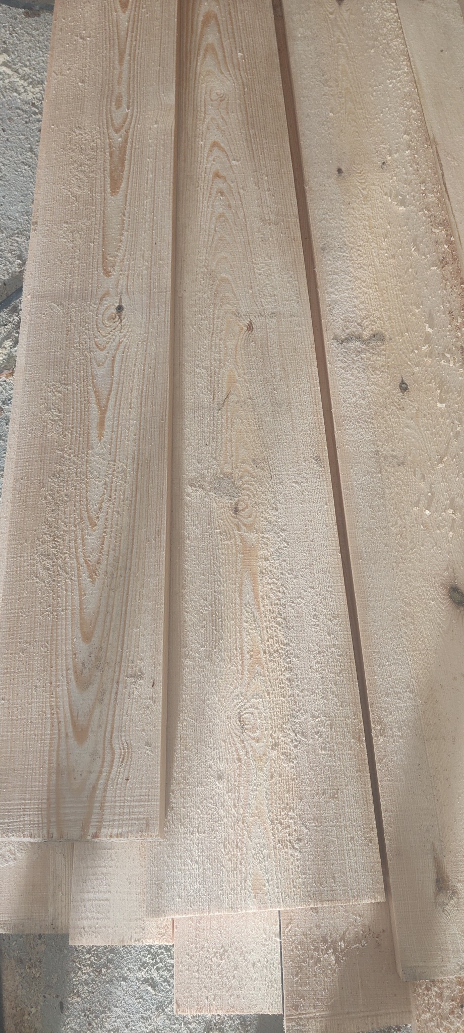Surowe, jasne deski z widocznym usłojeniem i śladami obróbki, ułożone warstwami na szarej, betonowej powierzchni. Widoczne drobne wióry i pył drzewny.