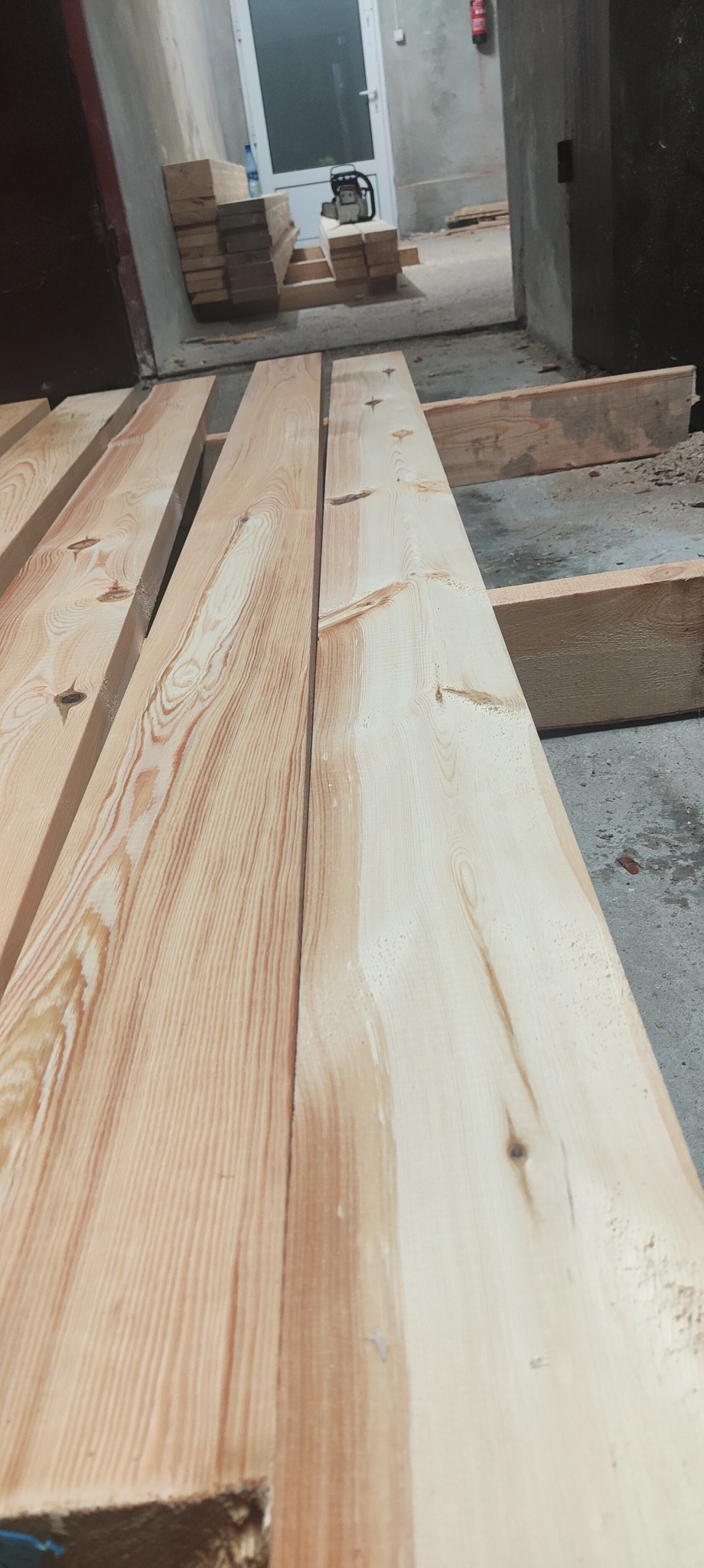 Surowe, jasne deski sosnowe ułożone w stos, z widocznym wzorem słojów i małymi sękami, w tle drzwi i piła łańcuchowa.