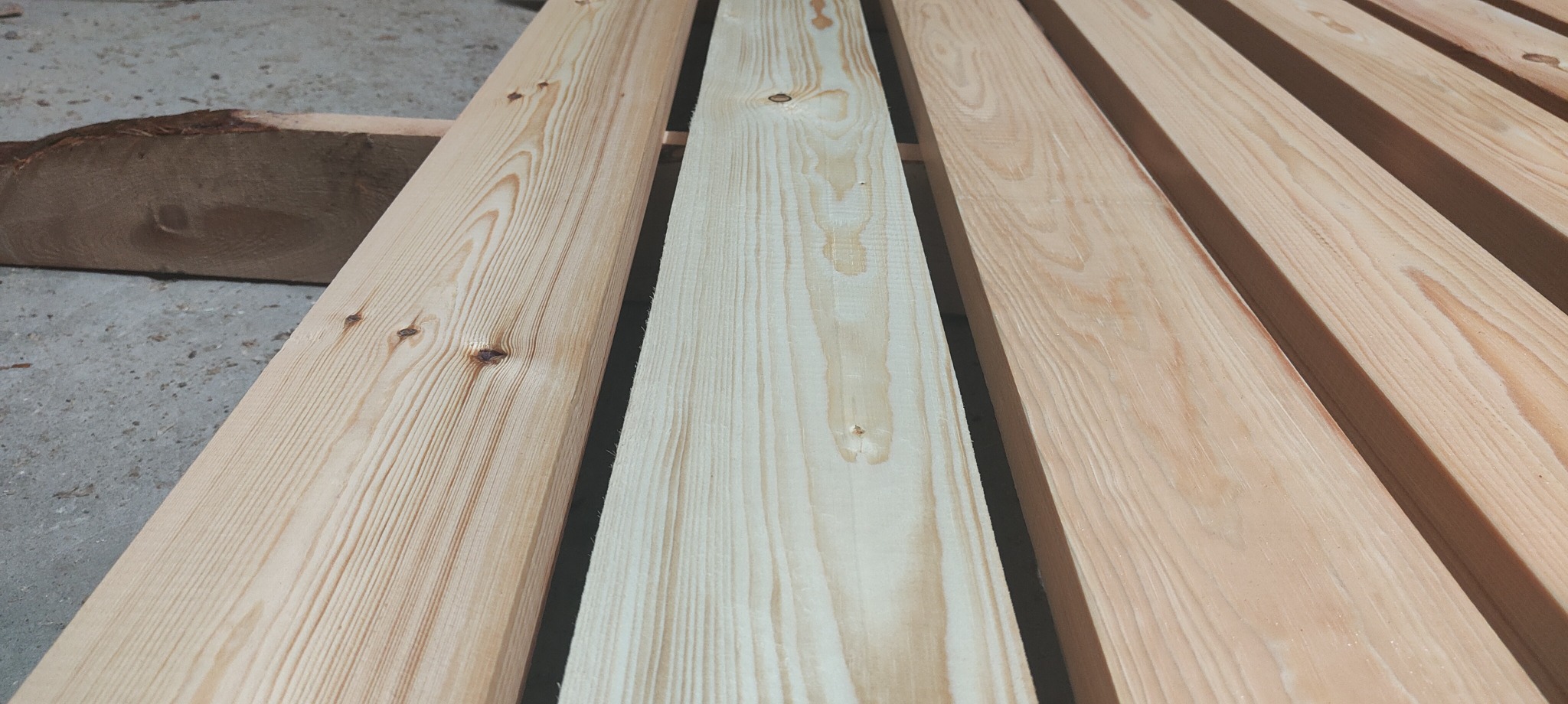 Ułożone deski sosnowe o różnym odcieniu, widoczne sęki i usłojenie drewna, fragment kory na boku, ułożone równolegle z widocznymi przerwami.