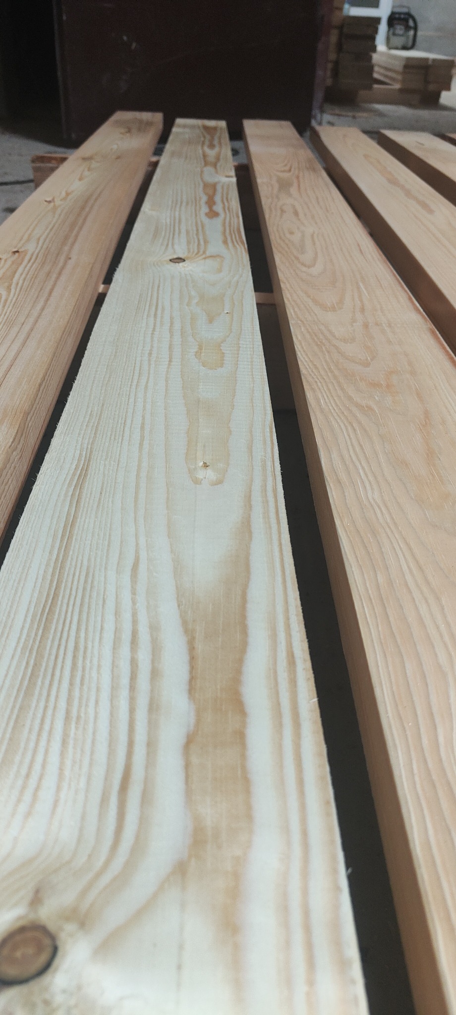 Ujęcie z bliska na ułożone równolegle, jasne deski z wyraźnym usłojeniem, oddzielone ciemnymi przekładkami, z lekko rozmytym tłem i stosem desek w tle.