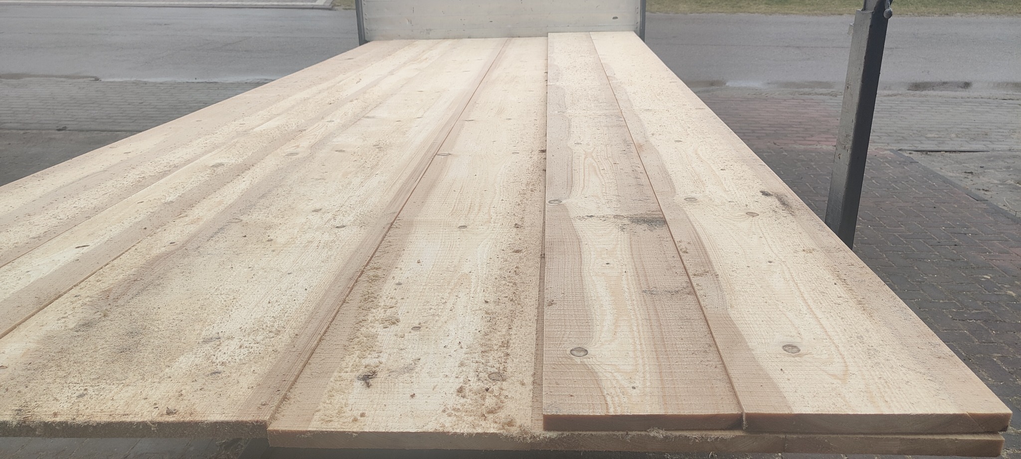 Świeżo przetarte, jasne deski ułożone równolegle, widoczne ślady trocin, transport na przyczepie lub w przestrzeni ładunkowej, szara kostka brukowa w tle.