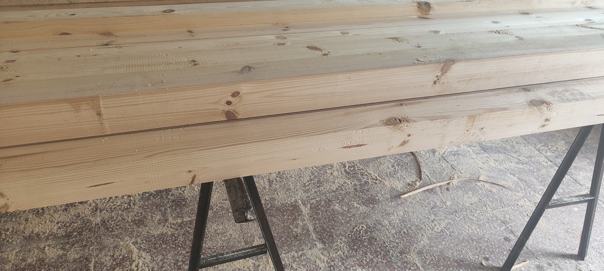 Ułożone kantówki z jasnego drewna iglastego, widoczne sęki i delikatne ślady po obróbce, leżące na metalowych kozłach w warsztacie w Rębielicach Szlacheckich, otoczone drobnymi wiórami.