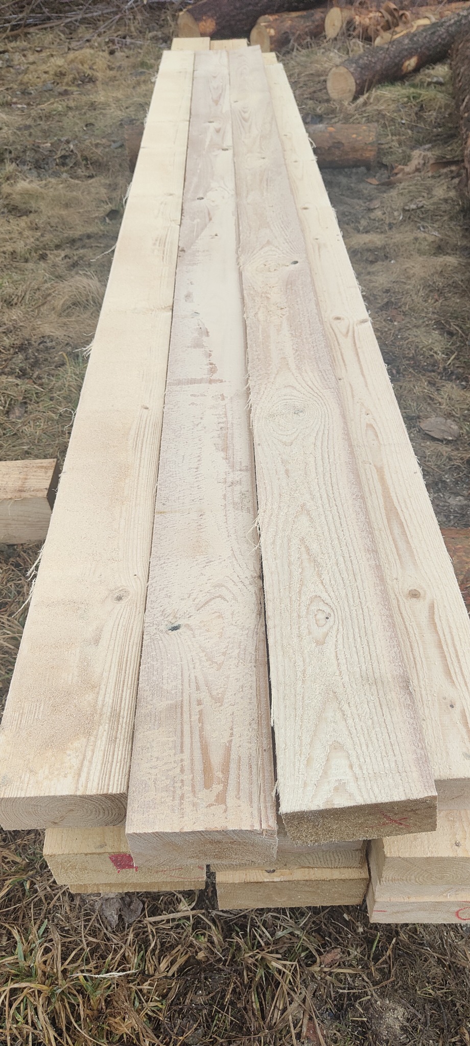 Stos świeżo przetartej, jasnej tarcicy iglastej ułożony na ziemi, widoczne ślady obróbki i czerwone oznaczenia na niektórych elementach.