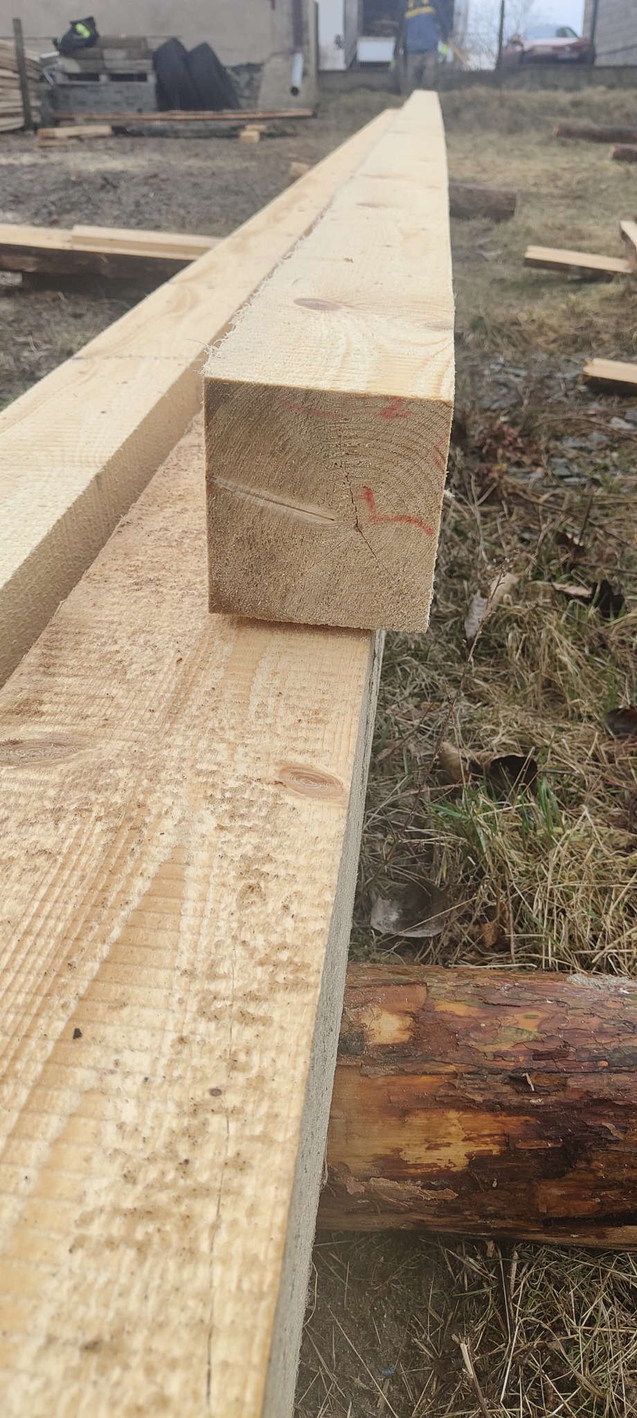 Kwadratowy przekrój świeżo obrobionej belki z jasnego drewna z czerwoną kreską pomiarową, ułożonej na długiej desce, widoczne sęki i struktura drewna, w tle trawa i budynek.