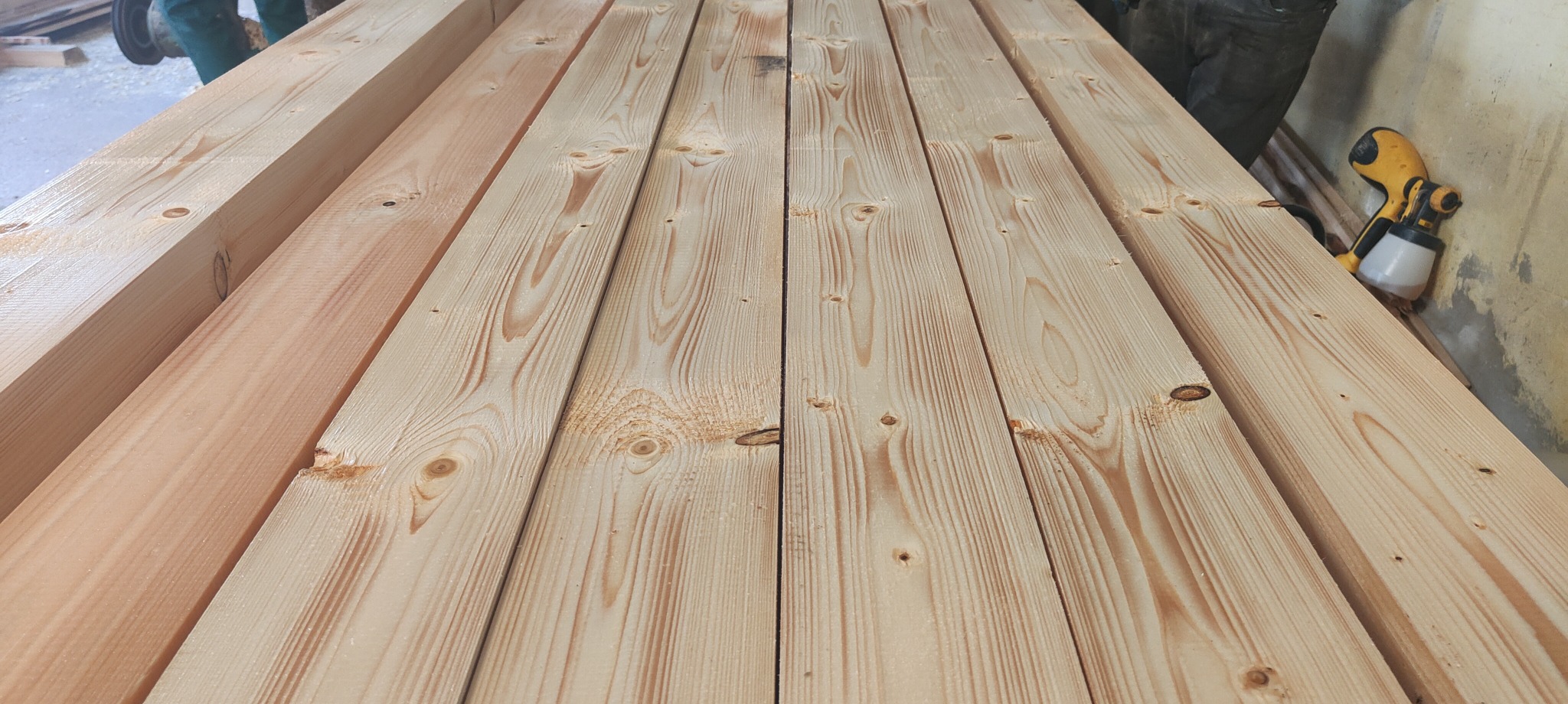 Stos jasnej, świeżo obrobionej tarcicy sosnowej, ułożonej równolegle, z widocznymi słojami i sękami, obok żółtego pistoletu do malowania.