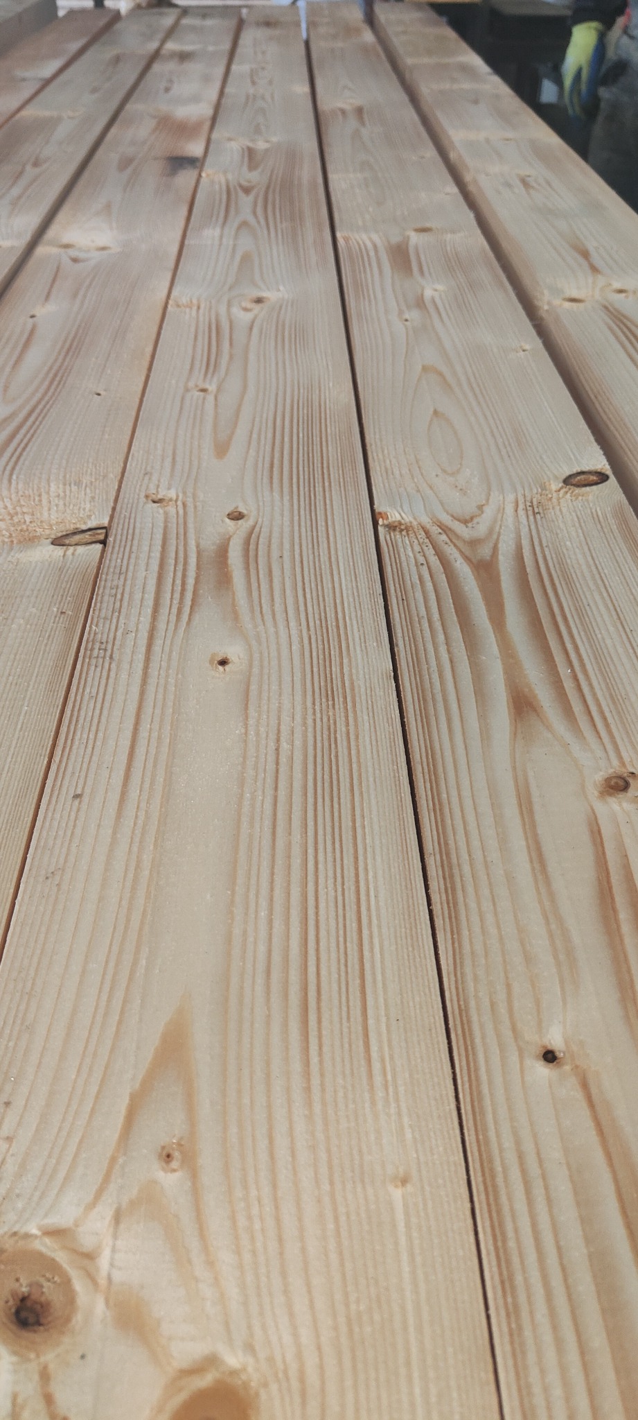 Stos jasnych, heblowanych desek sosnowych z widocznymi słojami i sękami, ułożonych równolegle w magazynie.