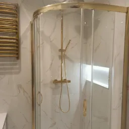 Wykończona kabina prysznicowa z złotymi elementami, dopasowana do marmurowych płytek. Widoczny grzejnik drabinkowy w kolorze złota i oświetlenie wnękowe.