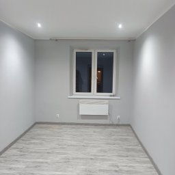 DARIUSZ TARNOWSKI GLADIATOR - Pusty pokój po remoncie: szare ściany, drewnopodobna podłoga, białe okno z grzejnikiem. Widoczne gniazdko i oświetlenie sufitowe.