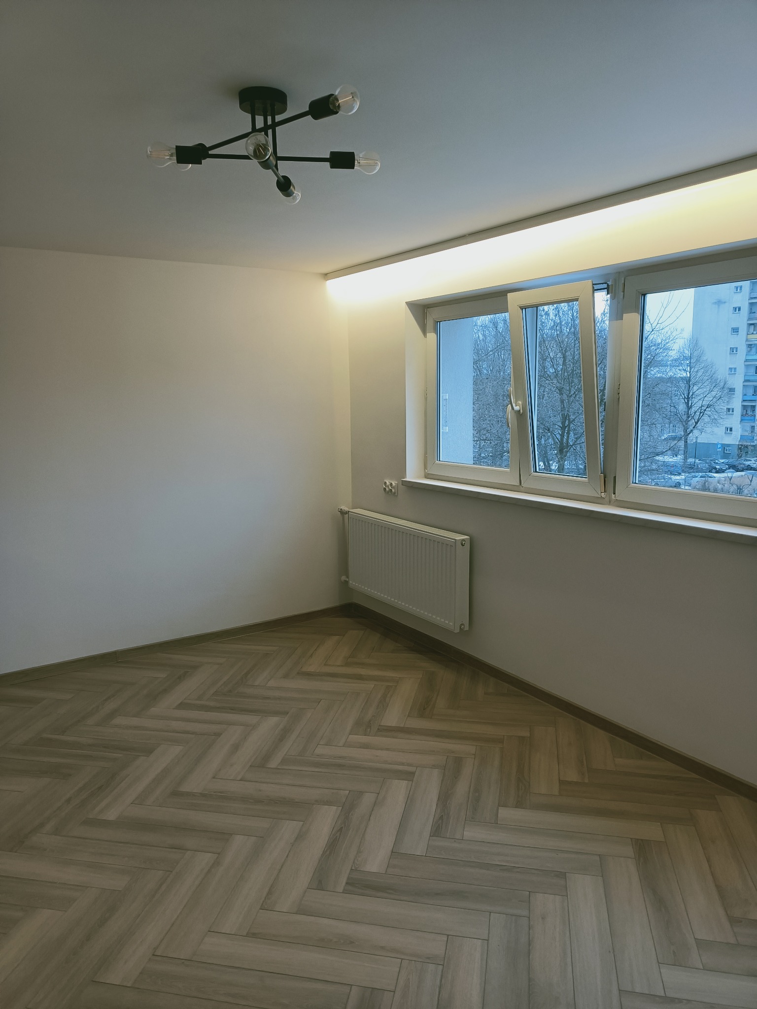 Pusty pokój po remoncie z parkietem ułożonym w jodełkę, białymi ścianami, grzejnikiem i nowoczesną lampą sufitową z żarówkami Edisona.