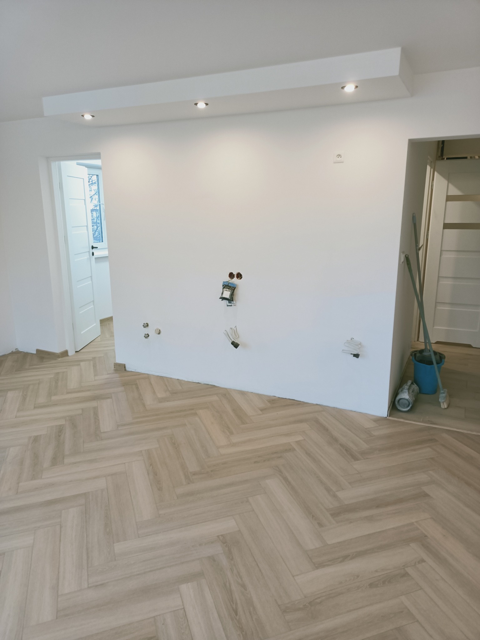 Nowe panele podłogowe ułożone w jodełkę, białe ściany z wyprowadzonymi instalacjami elektrycznymi i hydraulicznymi, widoczne narzędzia i wiadro, przygotowane do dalszych prac wykończeniowych.