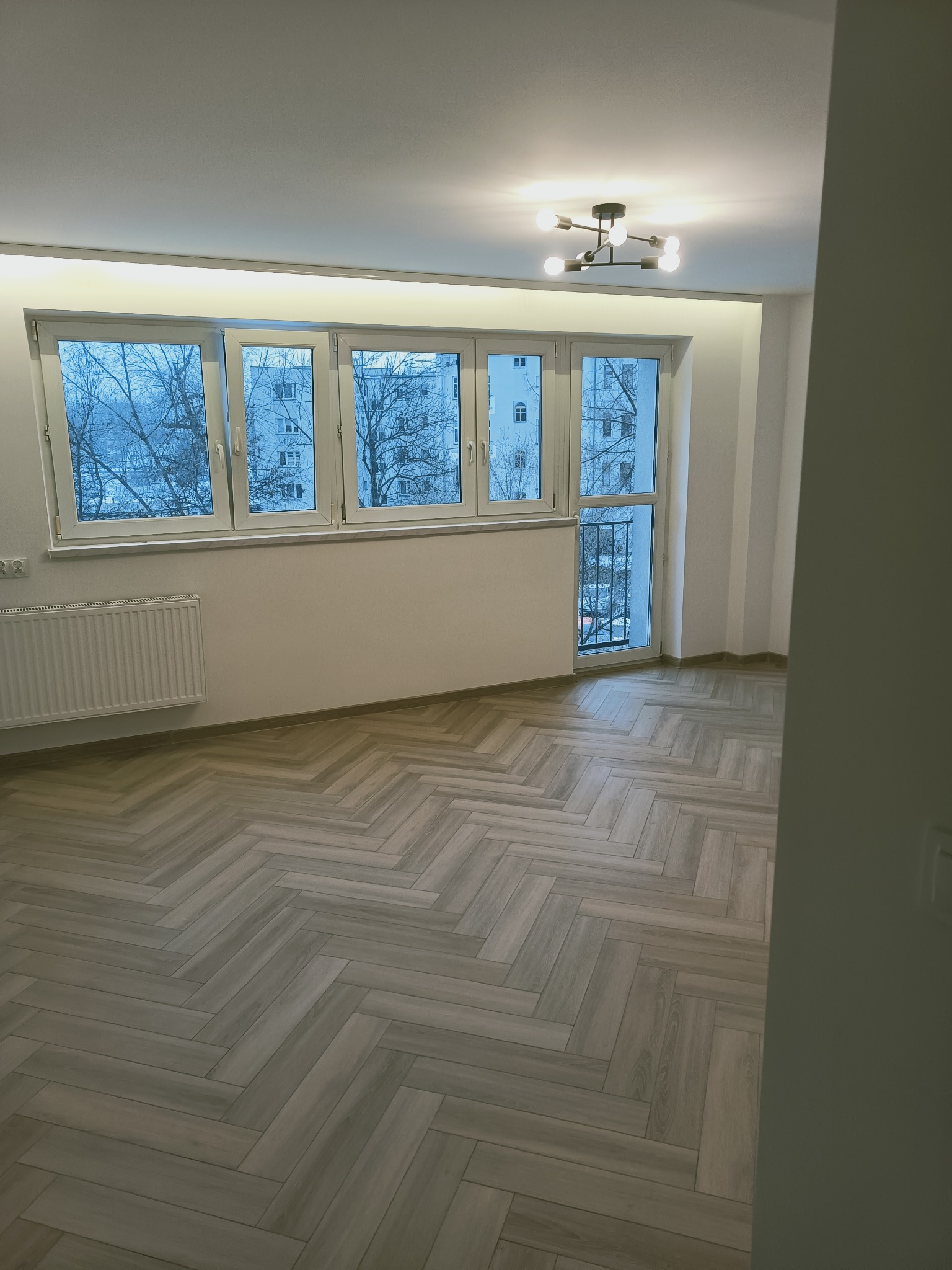 Pusty pokój po remoncie z parkietem ułożonym w jodełkę, białymi ścianami, grzejnikiem i oknem z widokiem na blok w zimowy dzień.