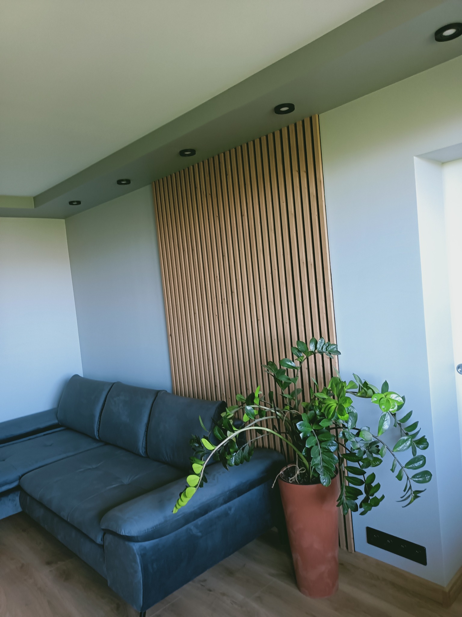 Szary narożnik z drewnianą ścianką działową w pionowe listwy; doniczka z zieloną rośliną. Nowoczesne wykończenie wnętrza, oświetlenie punktowe.