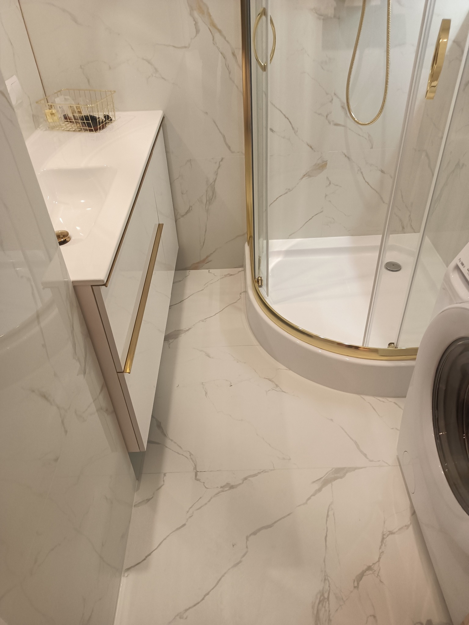 Elegancka łazienka po remoncie: białe marmurowe płytki, kabina prysznicowa ze złotymi detalami, biała szafka pod umywalką z złotymi uchwytami, pralka.