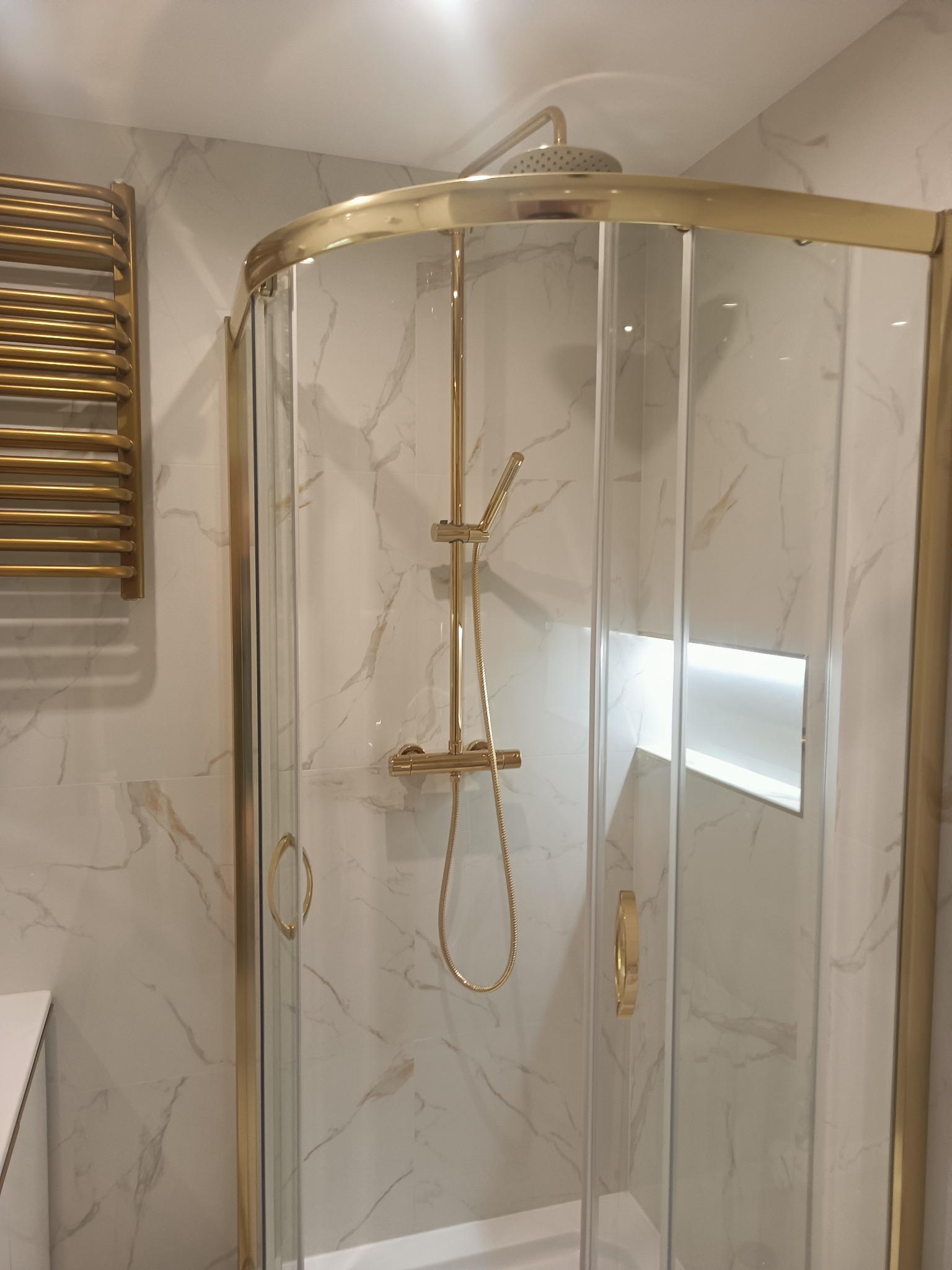 Wykończona kabina prysznicowa narożna z armaturą w kolorze złota, na tle płytek imitujących marmur. Widoczny grzejnik drabinkowy w tym samym odcieniu.