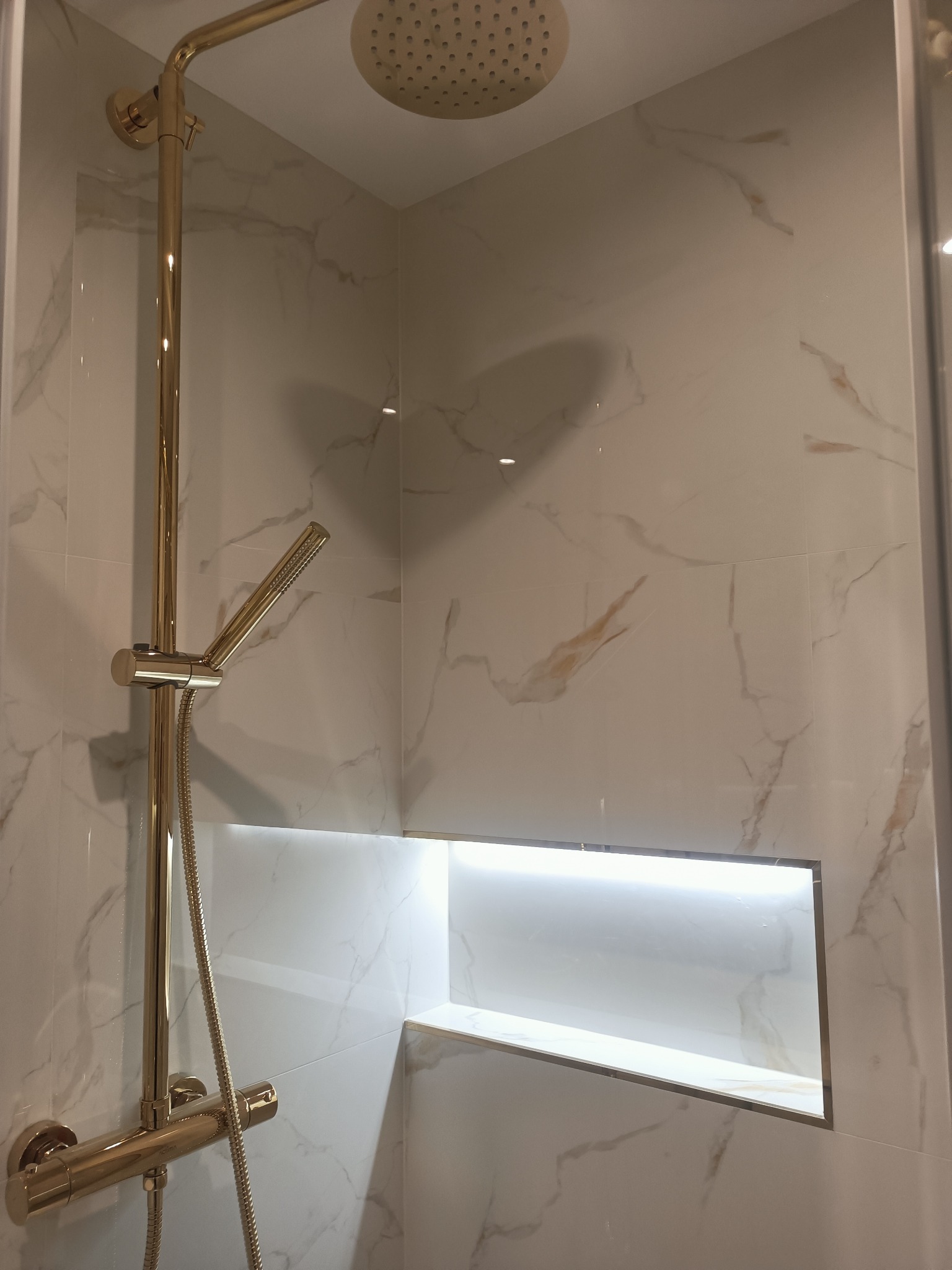 Elegancka kabina prysznicowa z marmurowymi płytkami i złotą armaturą. W ścianie wnęka z podświetleniem LED. Nowoczesny design łazienki.