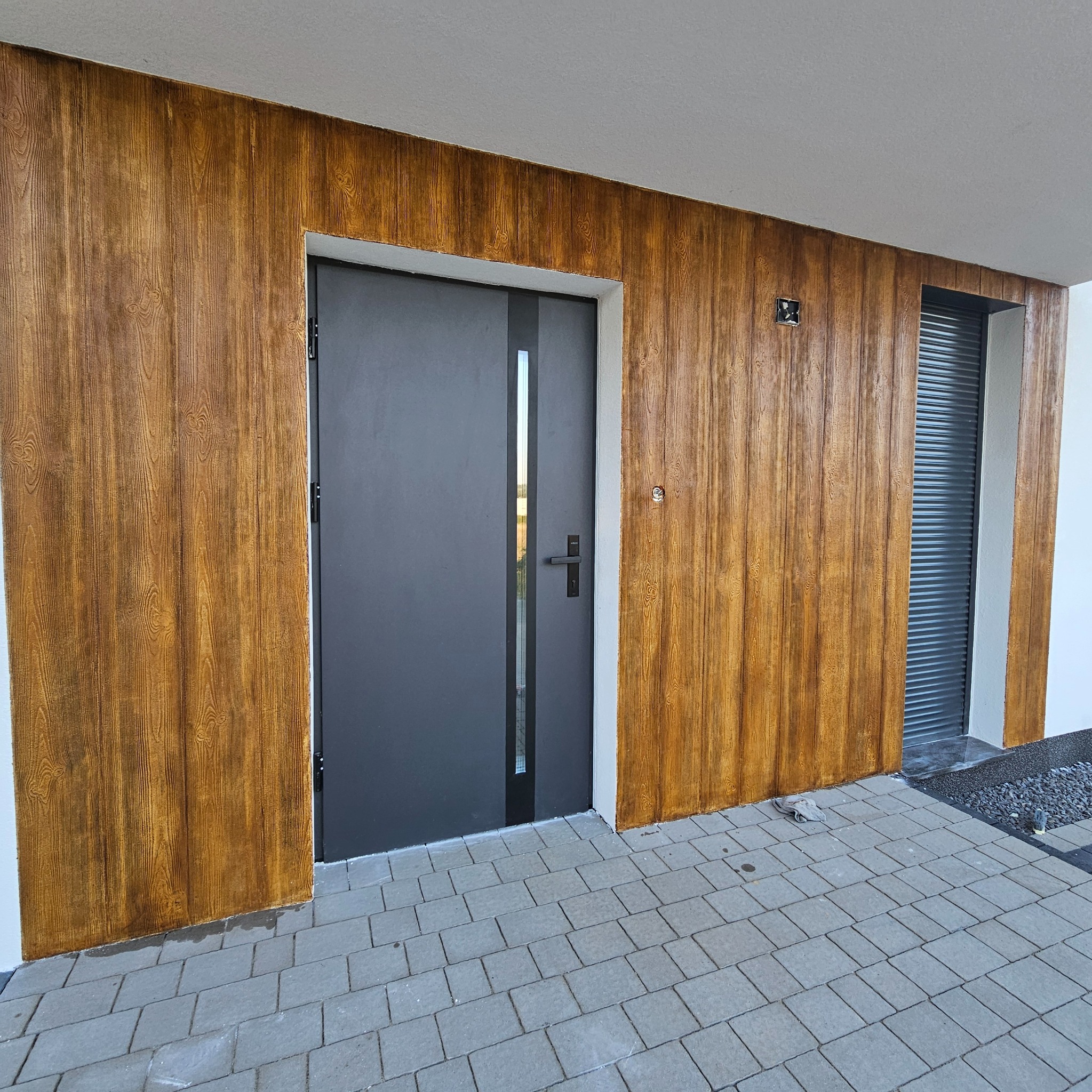 Elewacja domu z tynkiem strukturalnym imitującym drewno, szara nowoczesna stolarka drzwiowa i okienna, oraz kostka brukowa na podjeździe.