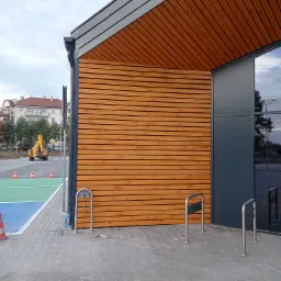 Elewacja budynku handlowego z drewnianą okładziną w poziomym układzie, szara blacha na dachu, widoczne stojaki rowerowe z logo Netto, w tle plac budowy z koparką.