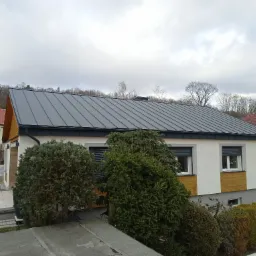 Nowoczesny dom jednorodzinny z szarym, panelowym dachem, widoczny z góry, z bujną zielenią w ogrodzie i pochmurnym niebem w tle.