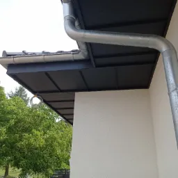 Zamontowana, srebrna rynna spustowa przy ścianie budynku z czarnym podbitką dachową i widocznym fragmentem dachu krytego blachą trapezową, z zielonym drzewem w tle.