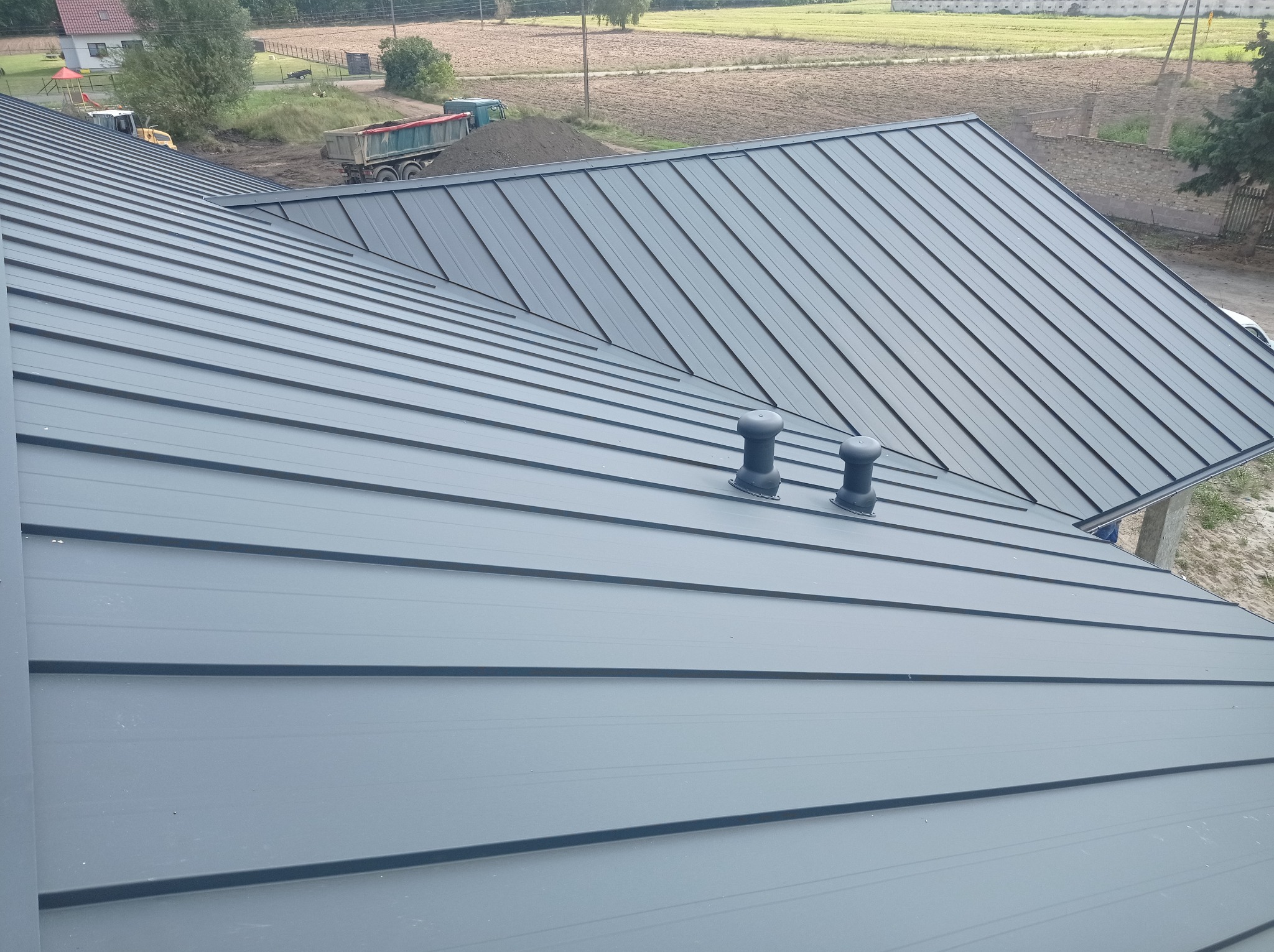 Szary dach z blachy na rąbek stojący, widoczne dwa kominki wentylacyjne, w tle plac budowy z koparką i wywrotką.