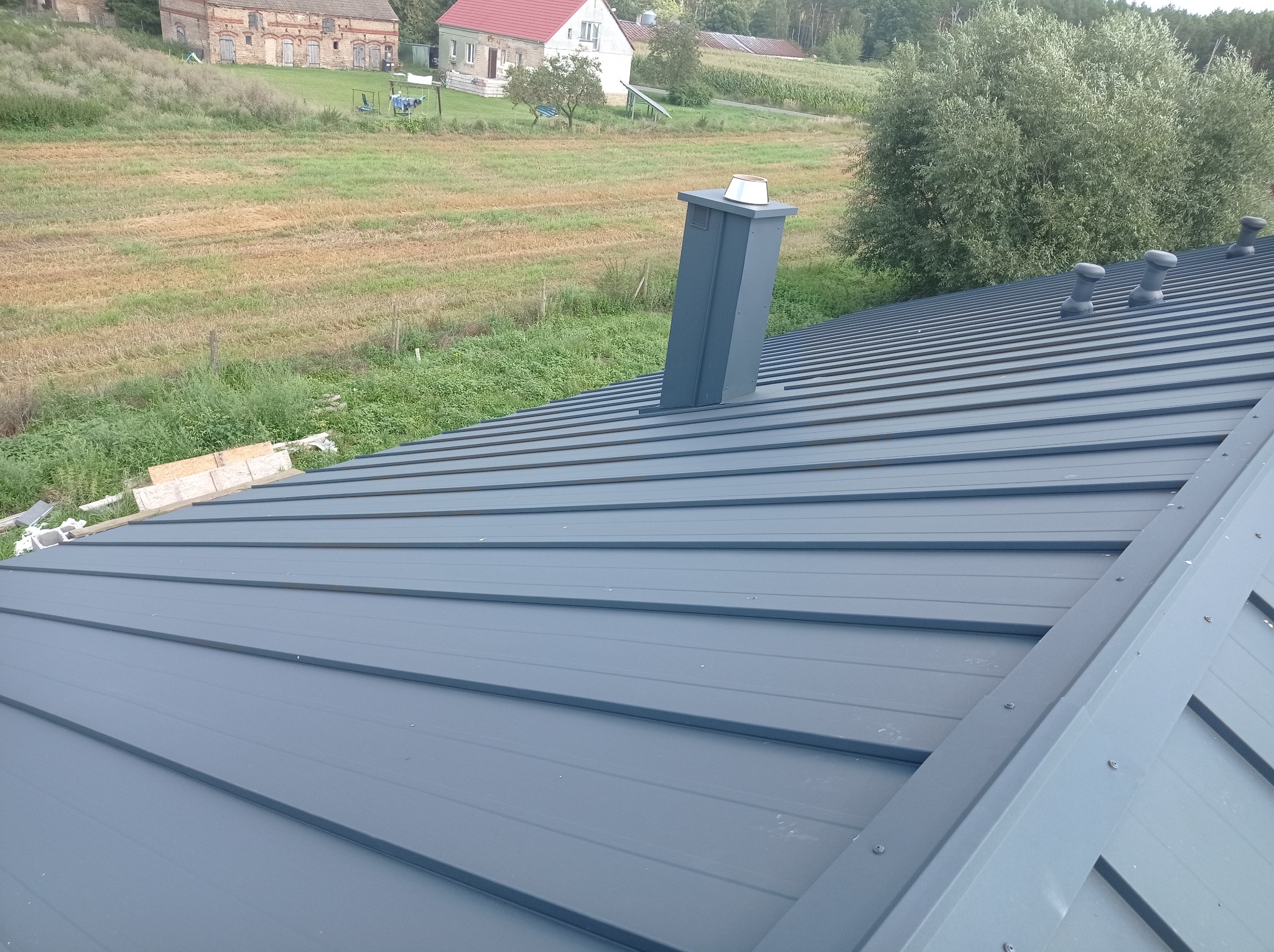 Nowy, szary dach z blachy trapezowej z kominem i odpowietrznikami w Gorzowie Wielkopolskim, na tle pola i zabudowań gospodarczych.