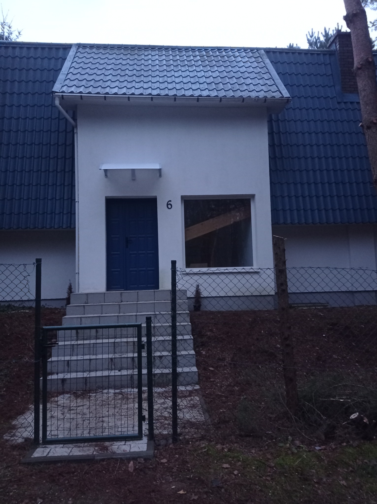 Front budynku z ciemnoniebieskim dachem z dachówki, niebieskimi drzwiami wejściowymi i schodami, widziany przez ogrodzenie z siatki.