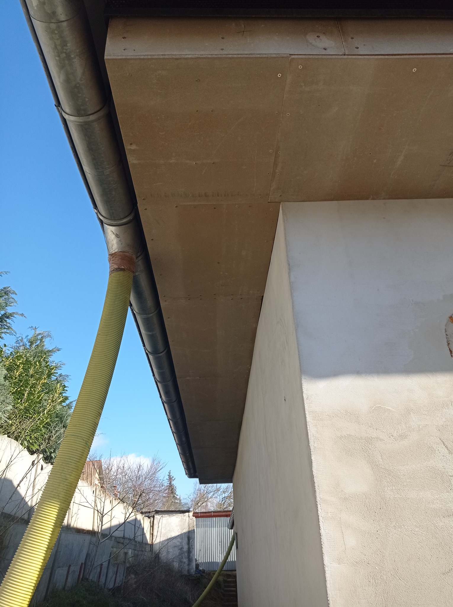 Metalowa rynna spustowa połączona żółtym, karbowanym wężem, widoczna na tle błękitnego nieba i fragmentu budynku z ocieplonym okapem.