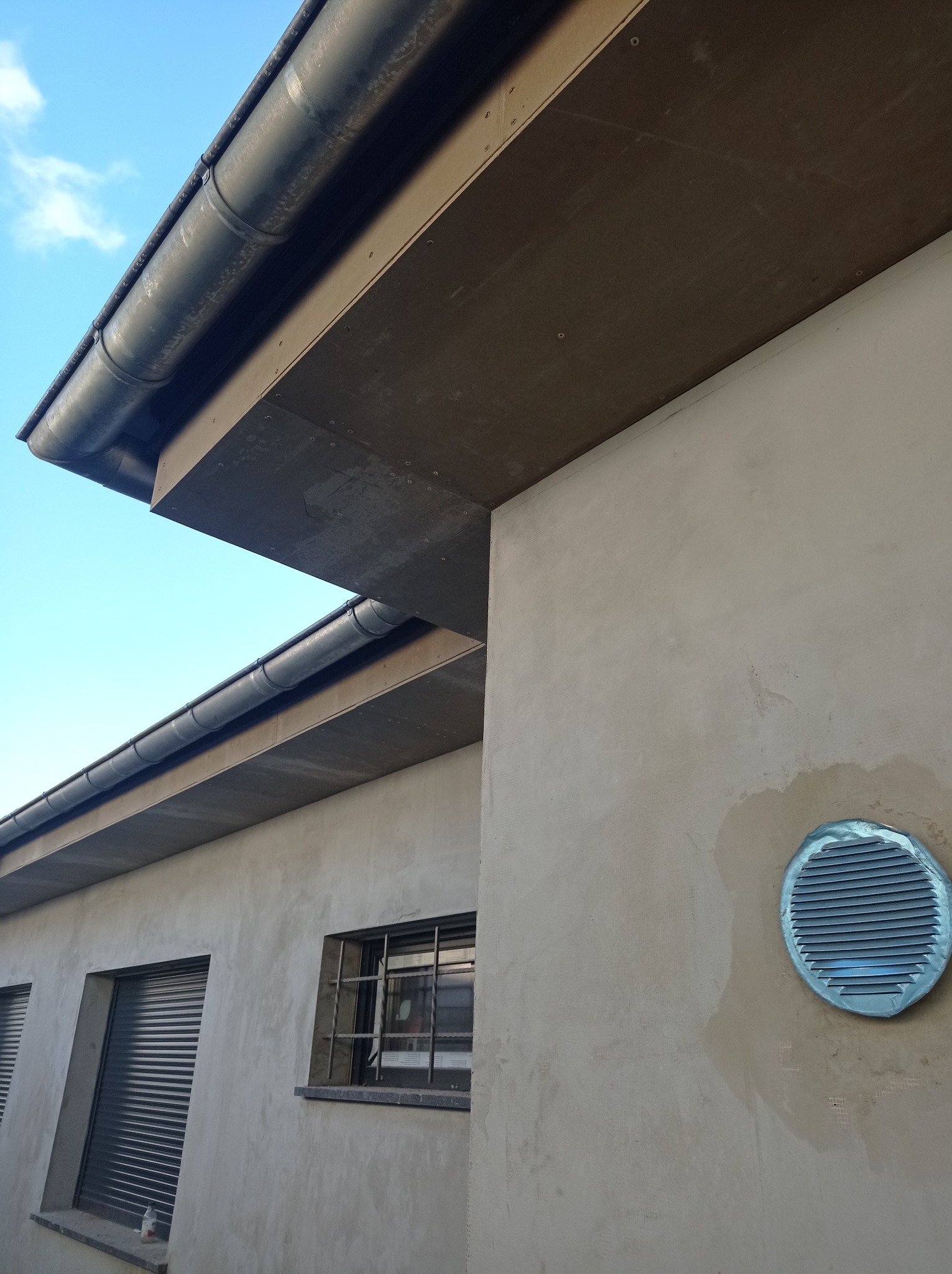 Szare rynny zamocowane na krawędzi dachu budynku z jasną elewacją, widoczny fragment nieba i okno z kratą.