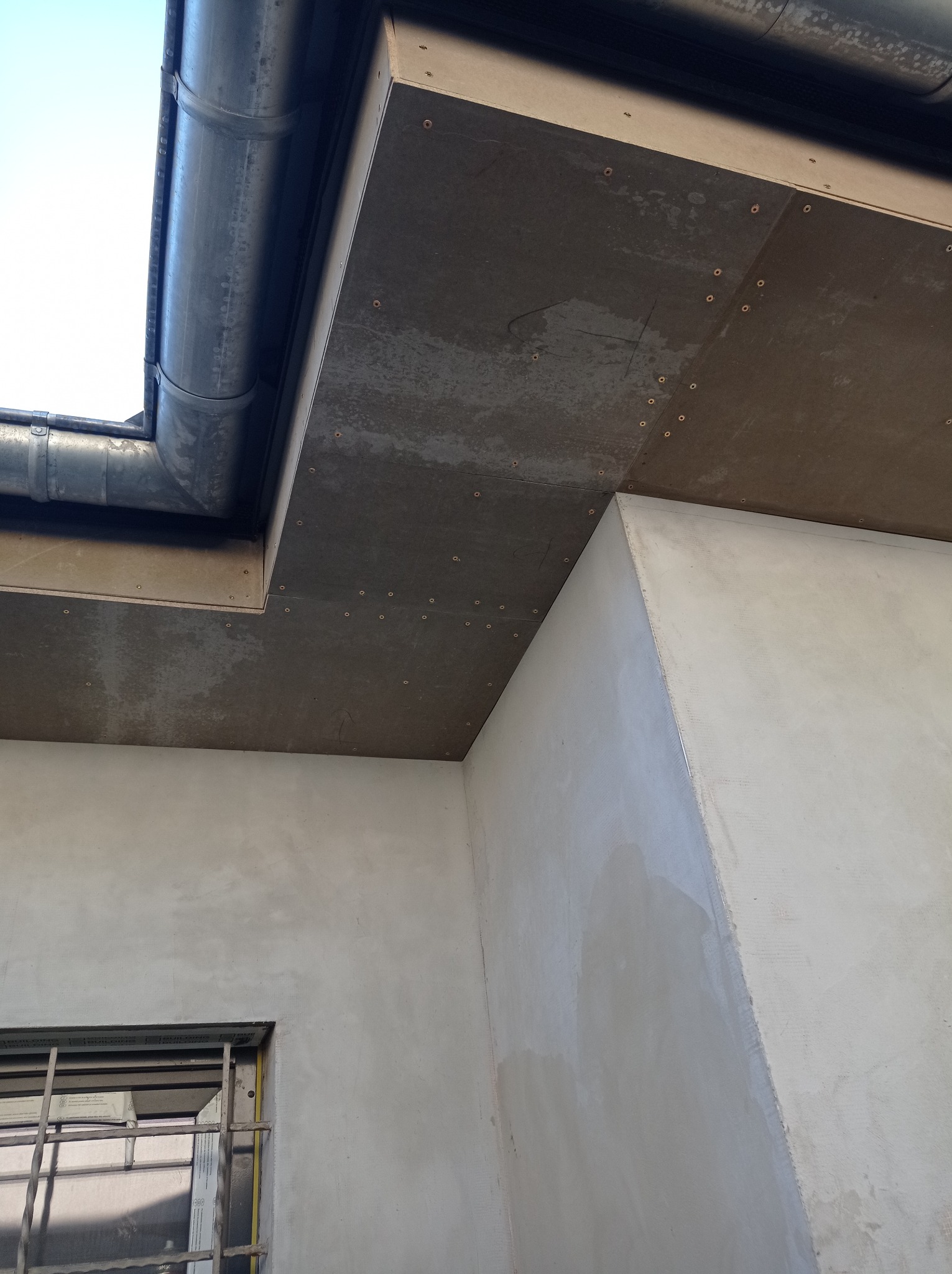 Fragment dachu z zamontowaną rynną, widoczne łączenia i mocowania, ściana budynku w tle.