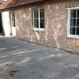 Fragment ściany budynku z cegły, z oknami w białych ramach, widoczny wykop wzdłuż fundamentu, biały samochód dostawczy w tle oraz worki z materiałami budowlanymi.