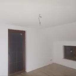 Poddasze po świeżym malowaniu, widoczne białe ściany, drewniane okno oraz drzwi z szybami, surowa żarówka na kablu zwisająca z sufitu.