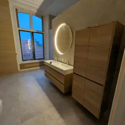 Nowoczesna łazienka z drewnianymi szafkami pod umywalką, okrągłym lustrem z podświetleniem LED i szarą podłogą z płytek, widok na budynki przez duże okno.