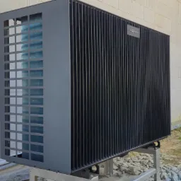 Czarna pompa ciepła Viessmann na zewnątrz budynku z białego pustaka, widoczna z boku, na metalowej konstrukcji, z elementami instalacji i kamieniami na ziemi.