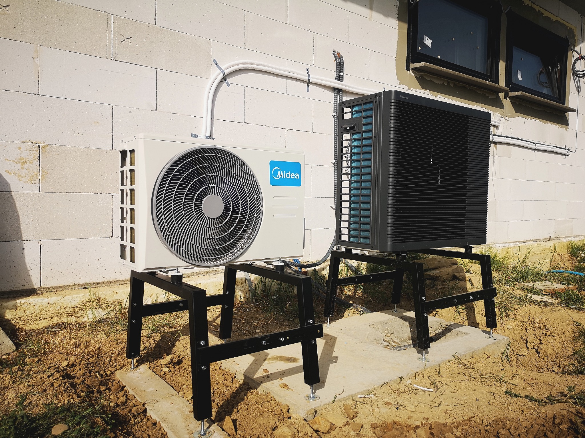 Dwa klimatyzatory typu split, Midea i Panasonic, zamontowane na metalowych podstawach na zewnątrz budynku z białego bloczka, z widocznymi przewodami instalacyjnymi.
