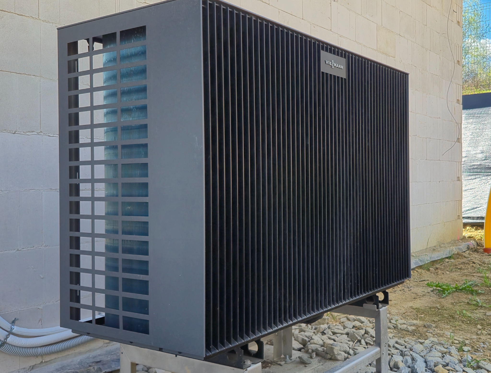 Czarna pompa ciepła Viessmann na zewnątrz budynku z białego pustaka, widoczna z boku, na metalowej konstrukcji, z elementami instalacji i kamieniami na ziemi.
