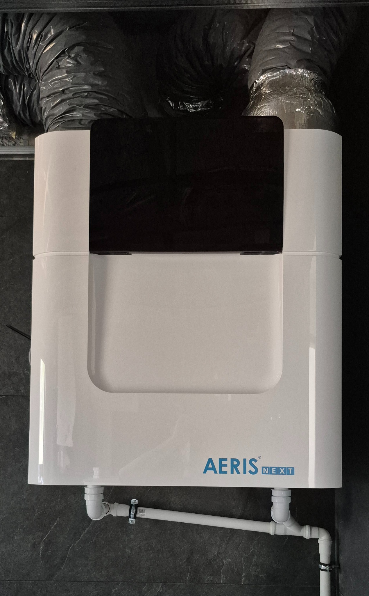 Biała jednostka rekuperacji Aeris Next z podłączonymi rurami, widoczna na tle szarej ściany i przewodów wentylacyjnych. Ujęcie z dołu.