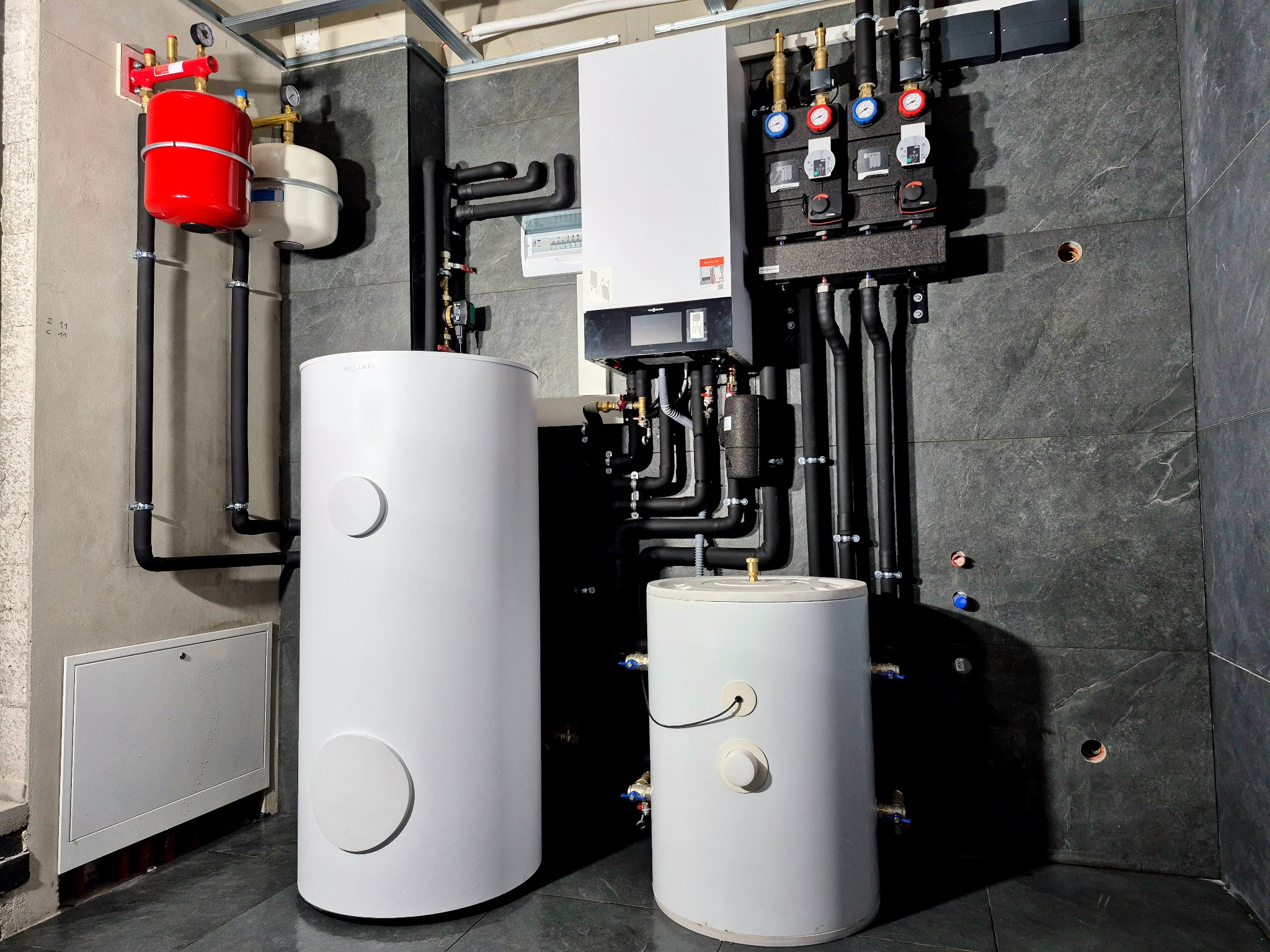 Kompletna instalacja grzewcza w kotłowni z piecem gazowym, zasobnikami wody, pompami i orurowaniem na tle szarej ściany z płytek imitujących kamień.