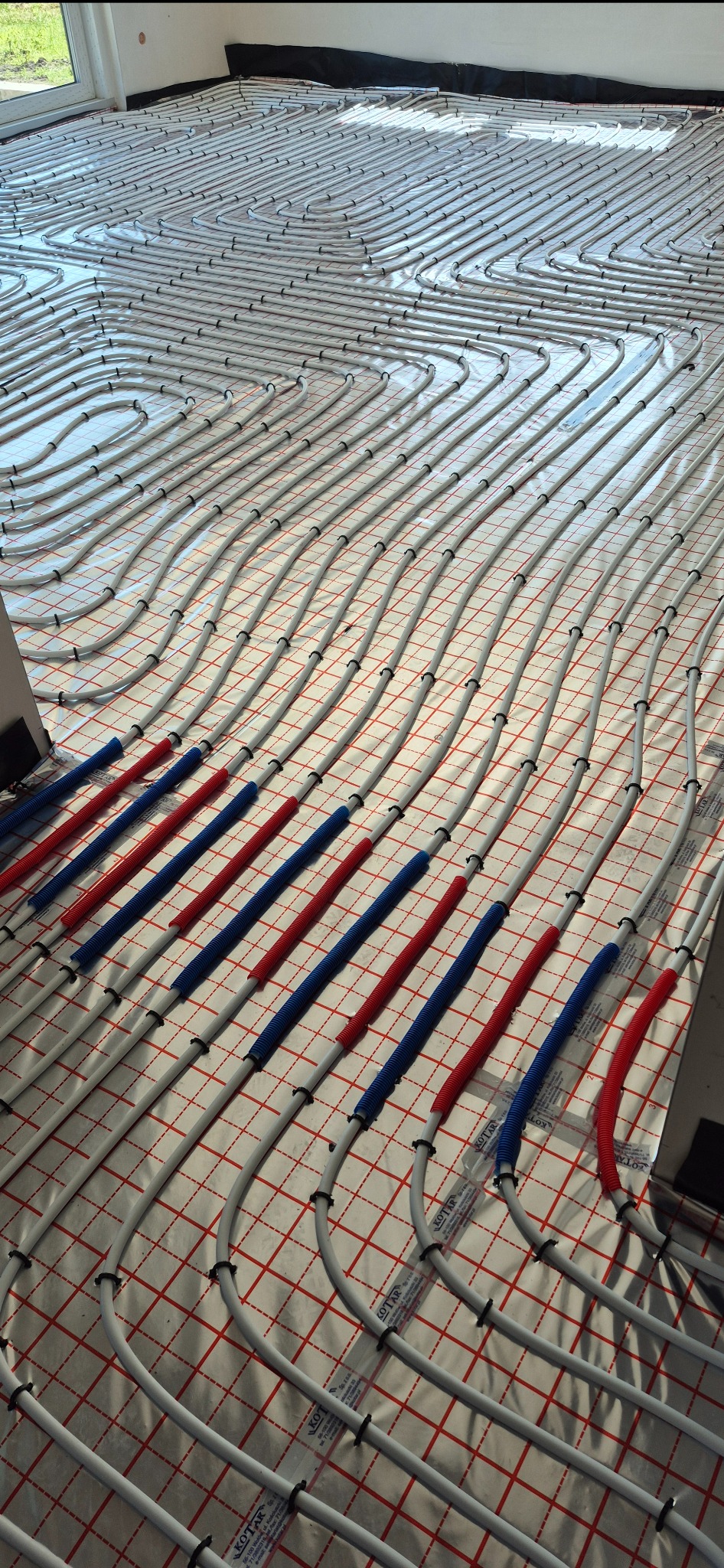 Ułożone białe rury ogrzewania podłogowego na folii z czerwoną siatką, połączone czerwonymi i niebieskimi rurami karbowanymi. Widok z góry.
