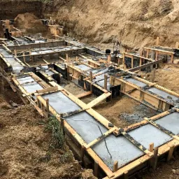 Wykop pod fundamenty z drewnianym szalunkiem i wylanym betonem, widoczne wystające pręty zbrojeniowe.