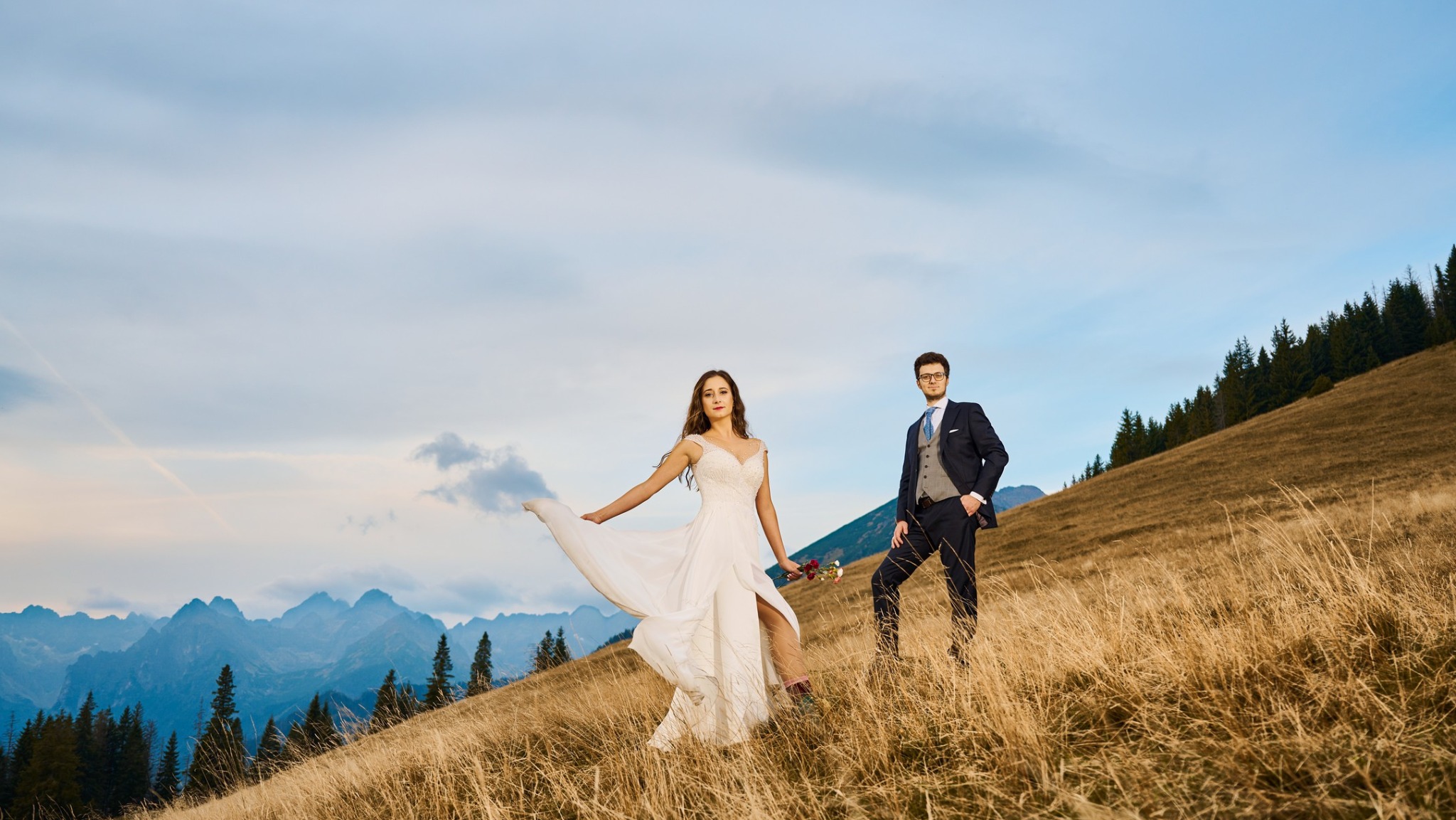 Para młoda: panna młoda w zwiewnej sukni z rozcięciem i bukietem, pan młody w garniturze, na tle górskiego krajobrazu w promieniach zachodzącego słońca.