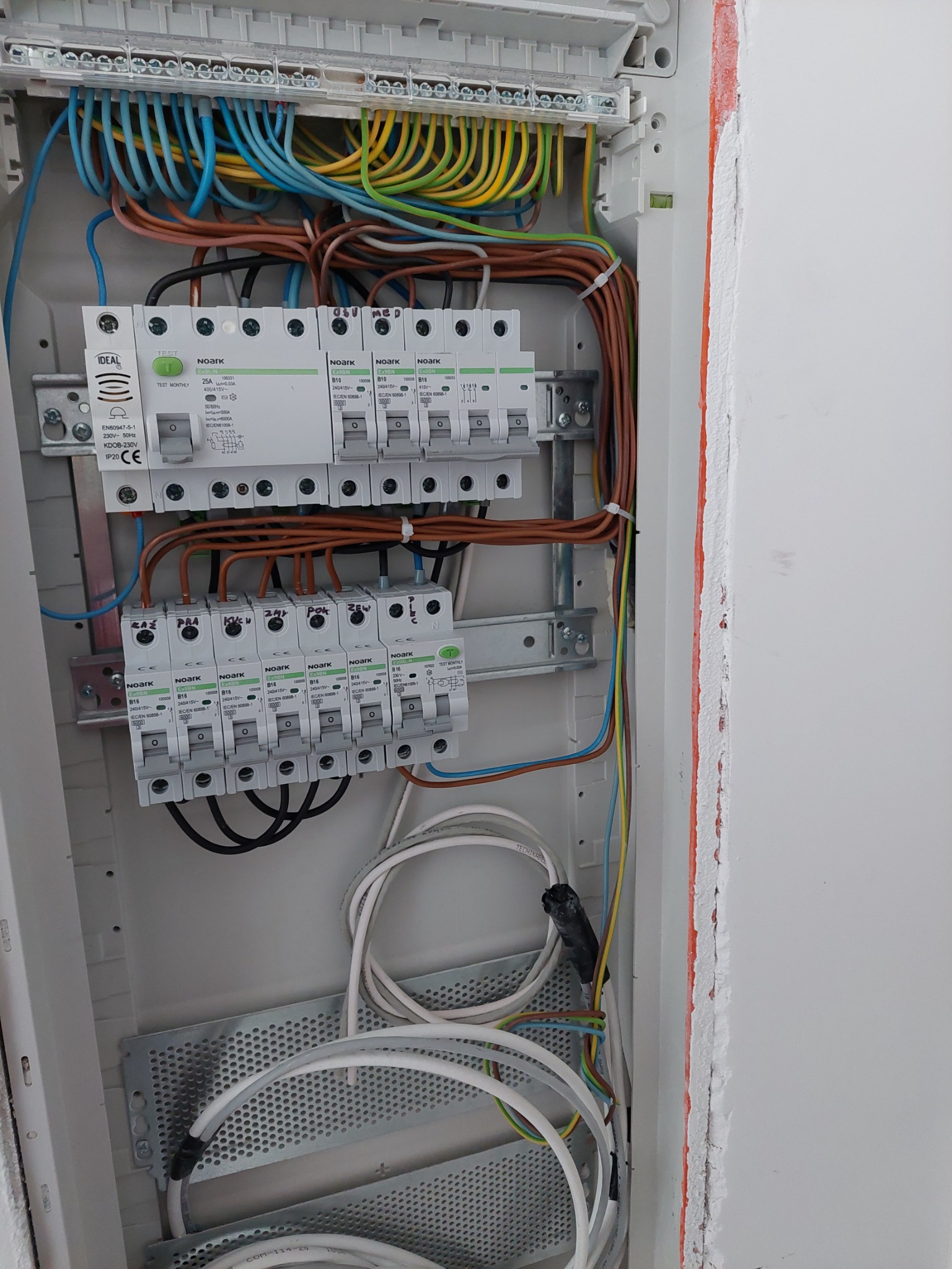 Otwarta skrzynka elektryczna z zainstalowanymi bezpiecznikami Noark, widoczne różnokolorowe przewody i szyny montażowe, instalacja w trakcie podłączania.