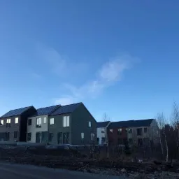 Kompleks kilku nowoczesnych domów w zabudowie szeregowej z drewnianą elewacją w różnych kolorach, na tle błękitnego nieba i lasu.