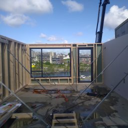 Marek Kwiecień - Wnętrze w trakcie budowy: drewniana konstrukcja szkieletowa z oknami, widok na miasto i rzekę, podpora stalowa, elementy mocujące, etap montażu ścian.